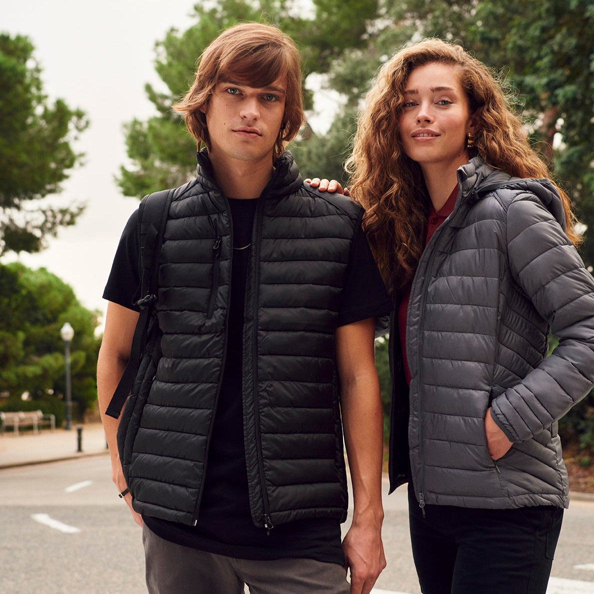 Two people wearing puffer vests standing on a street with trees in the background