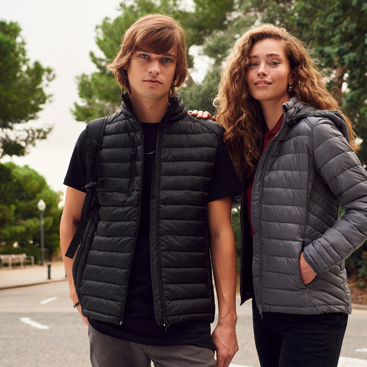 Two people wearing puffer vests standing on a street with trees in the background