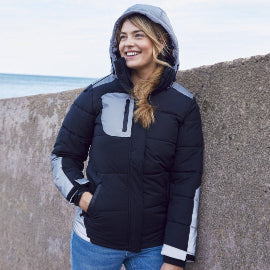 Person wearing a black and gray puffer jacket standing against a concrete wall with a beach in the background.