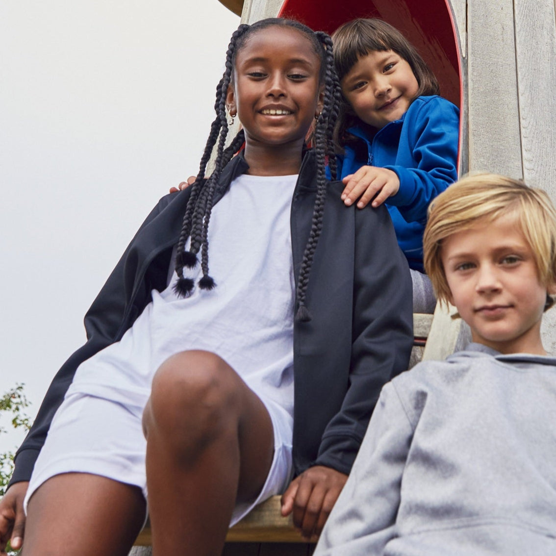 Three children sitting on a wooden playground structure with a clear sky background