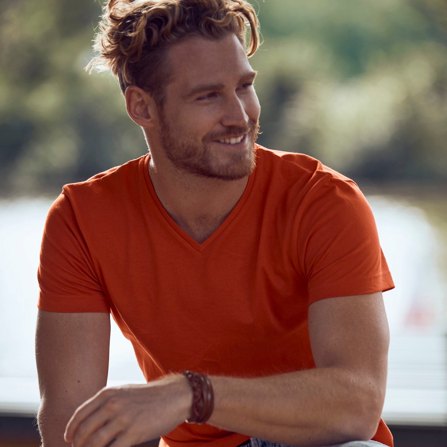 Man wearing an orange shirt and jeans sitting outdoors with a blurred background