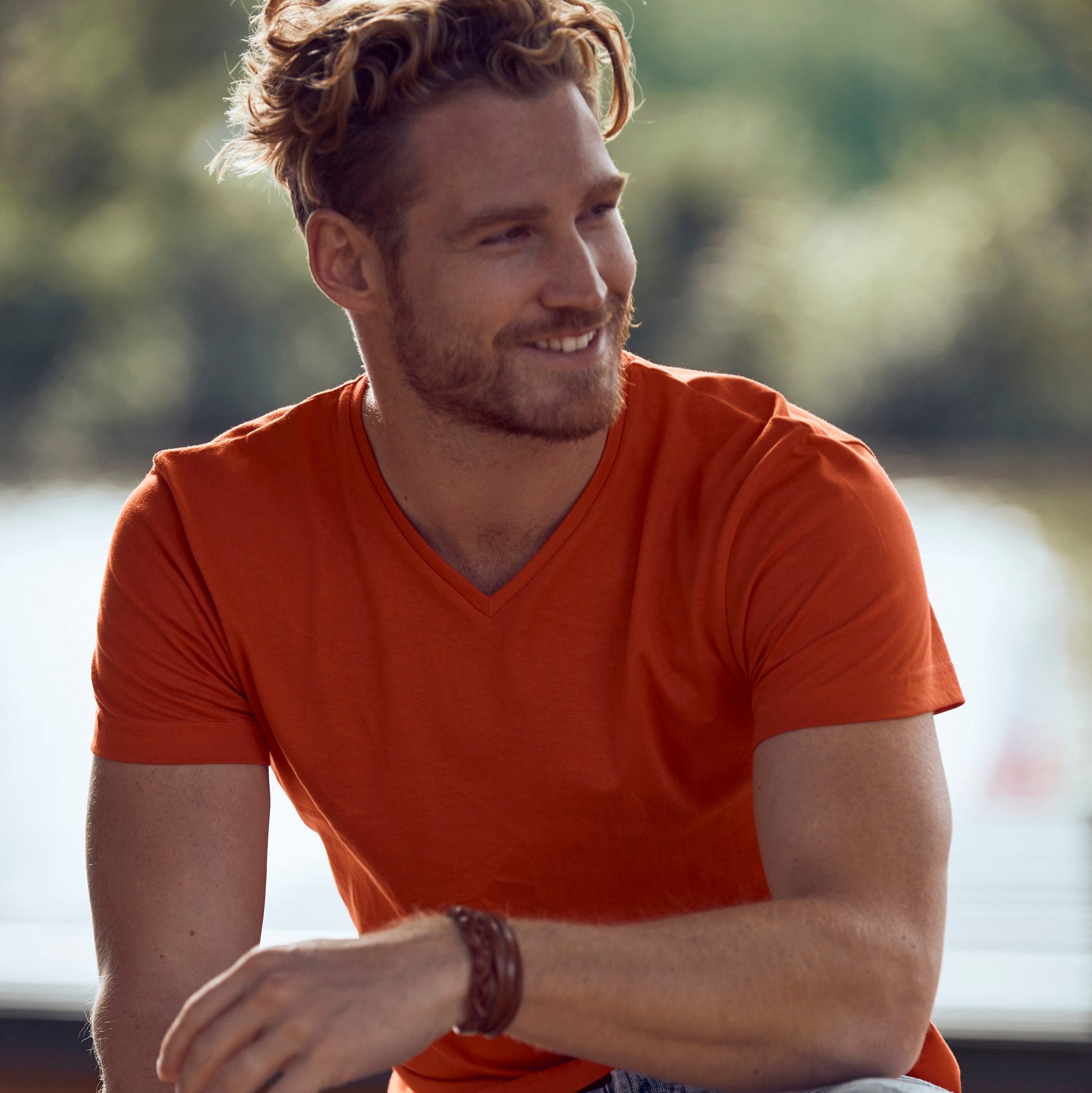 Man wearing an orange shirt and jeans sitting outdoors with a blurred background