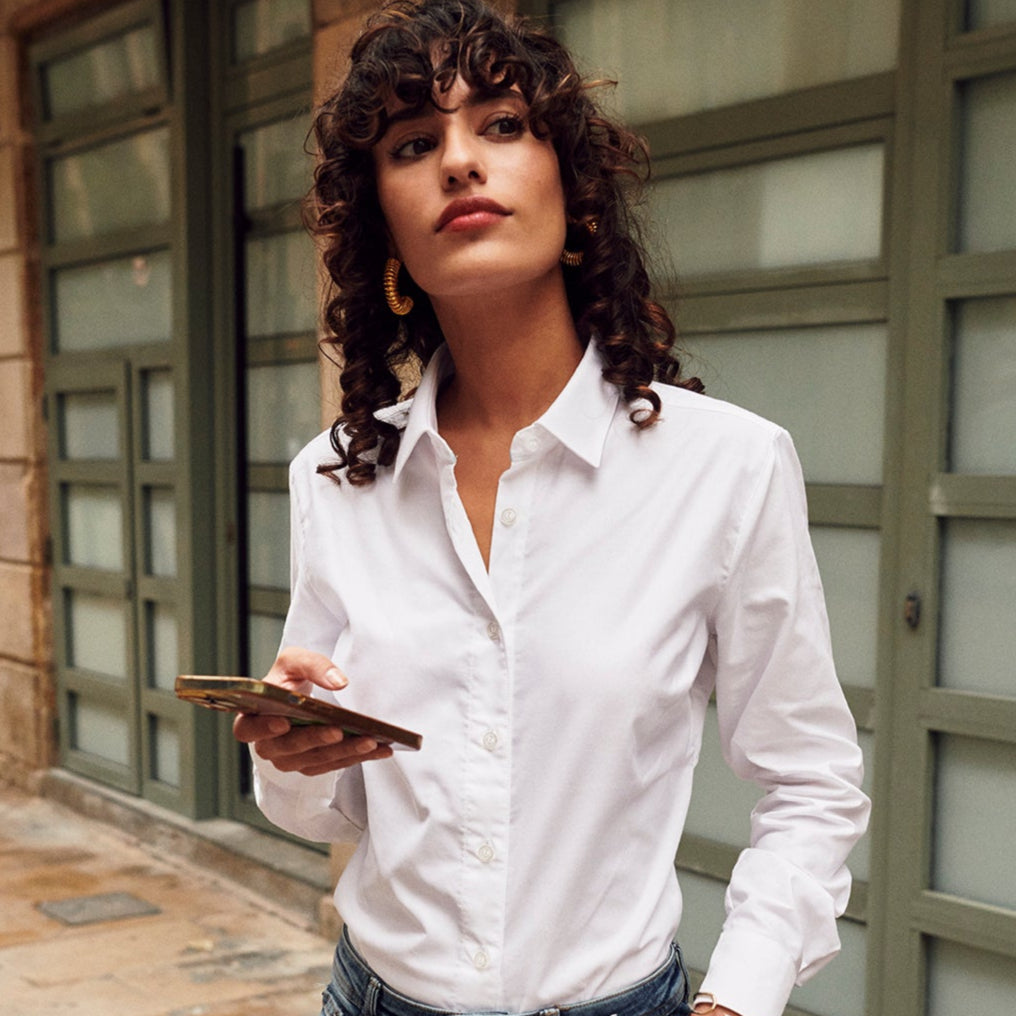 Woman in a white shirt and jeans holding a phone and a black bag on a sidewalk.