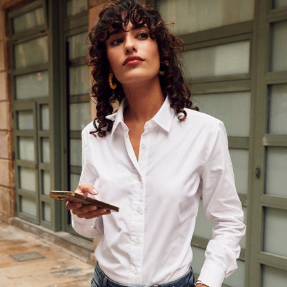 Woman in a white shirt and jeans holding a phone and a black bag on a sidewalk.