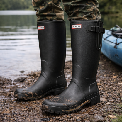 Black rubber boots worn by a person on a lakeside with camouflage pants and a kayak in the background.
